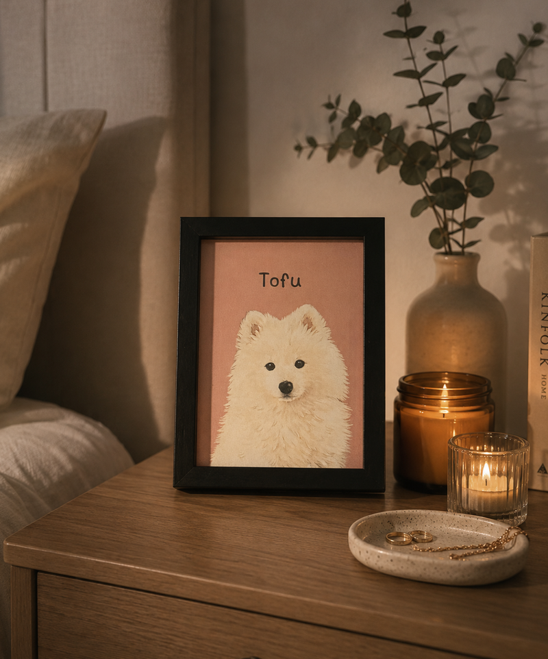 A framed art print featuring a white fluffy dog on a dusty pink background, styled on a wooden bedside table next to a lit candle, a ceramic vase with eucalyptus, and a small dish.