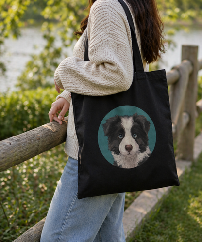 Person holding a black tote bag with a dog illustration, standing outdoors by a wooden fence.